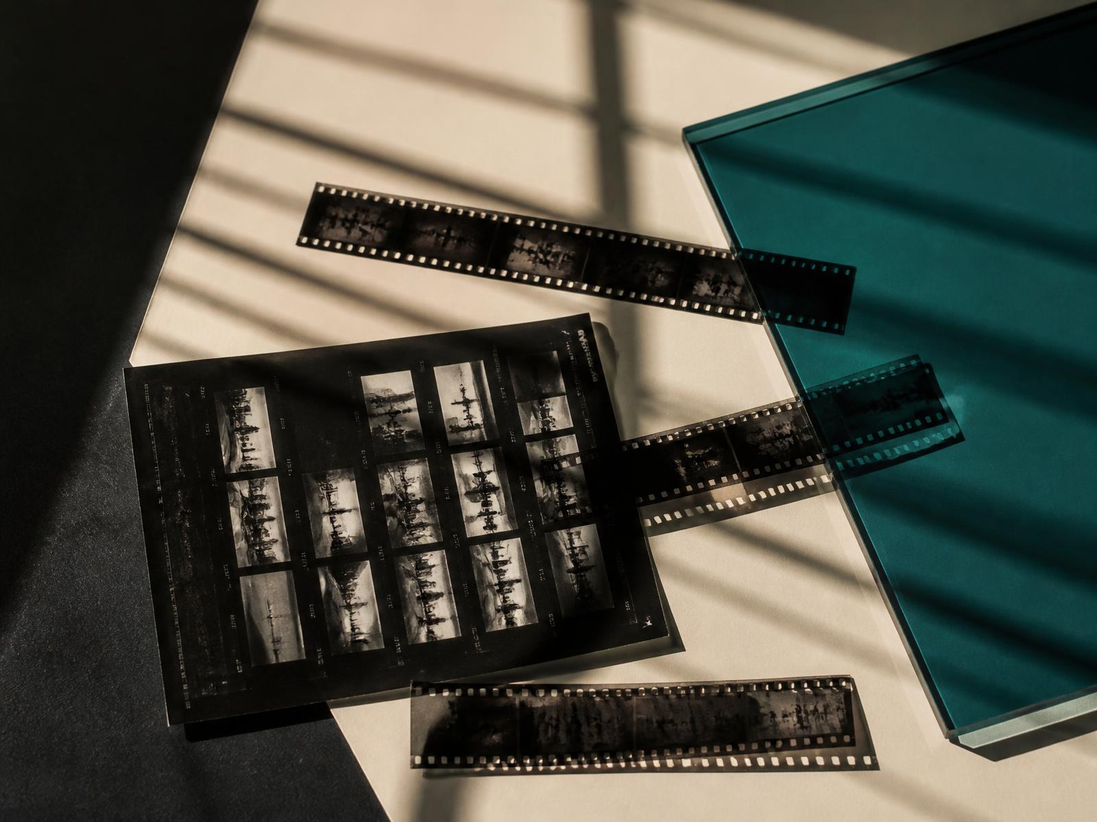 Film negatives and a contact sheet on cream paper, cut by hard studio light and a teal glass panel